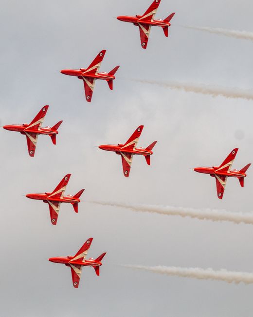 A formation of nine red aircrafts performing an synchronized aerial display against a cloudy sky during Farnborough Airshow week. The planes are flying in a diagonal pattern, leaving white smoke trail