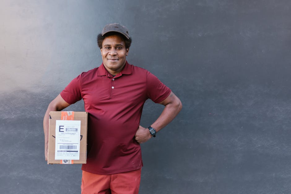 A smiling delivery person with dark curly hair, wearing a maroon polo shirt, matching maroon trousers, a dark cap, and a black wristwatch, stands against a plain dark wall. He holds a small cardboard 