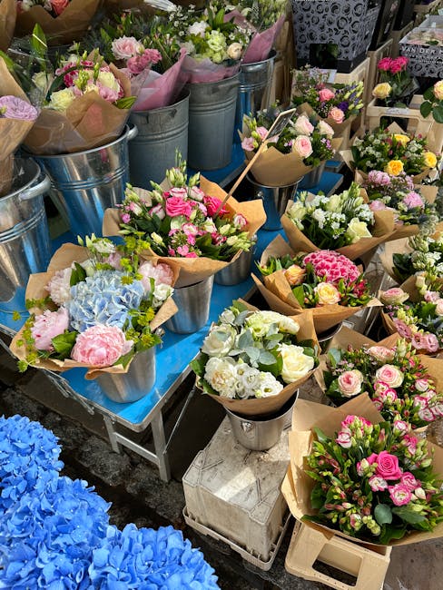 A vibrant display of freshly arranged floral bouquets positioned on a table outdoors, featuring a variety of flowers including pink roses, white carnations, pale yellow spray roses, and blue hydrangea