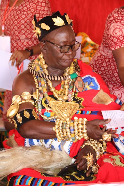 A woman dressed in vibrant traditional African attire, adorned with elaborate gold jewelry including necklaces, bracelets, and a crown, is seated indoors with a red and patterned background. She is ho