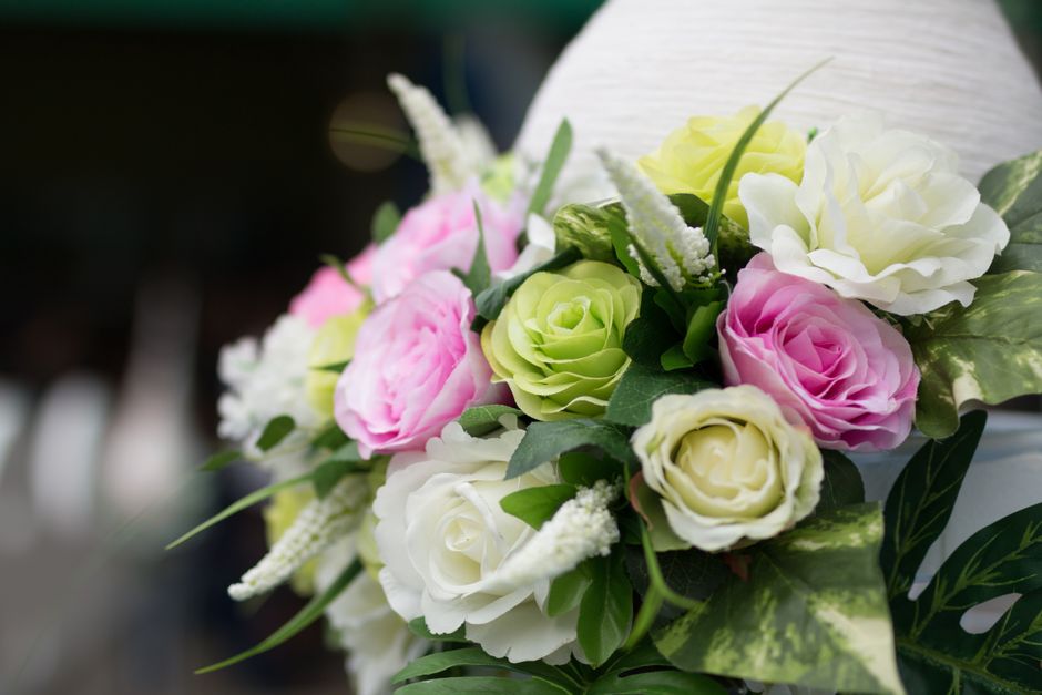 A close-up of a floral bouquet featuring cream, pink, and green roses, accented with white lavender-like blooms and various green leaves. The flowers are arranged in a rounded bouquet with fresh petal