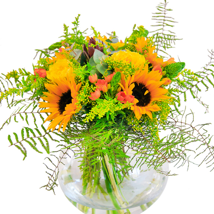From its first impression, this lavish arrangement delivers a flush of sunshine and warmth, reminiscent of a beautiful summer's day in Farnborough. Towering sunflowers showcase their large, honey-gold faces, radiating alongside plush, spiraled yellow garden roses that open with a soft, inviting elegance-just as the bloom of wildflowers might greet visitors exploring local spaces like Queen's Avenue. Shining among the yellow tones, vivid red clusters of Hypericum berries offer lively, eye-catching contrast, while airy ferns tumble along the bouquet's edge, mimicking the undulating greenery found just outside town. A careful scattering of golden tiny blossoms-likely solidago-adds a spark of texture, alongside slender green filler and the cool, distinct foliage of eucalyptus, which together introduce both depth and a hint of forest tranquility. The entire composition is presented in a rounded, glass vase, reminiscent of a fishbowl, with submerged stems visible through crystal water-a touch that enhances the arrangement's sense of openness and natural airiness. Bathed in natural light, the display feels both uplifting and serene, ideal for occasions when happiness deserves to be celebrated, like birthdays, anniversaries, or new beginnings in Farnborough. Every flower and leaf is arranged with the experienced hand of Flowers Farnborough, ensuring freshness and an unmistakeably local charm.