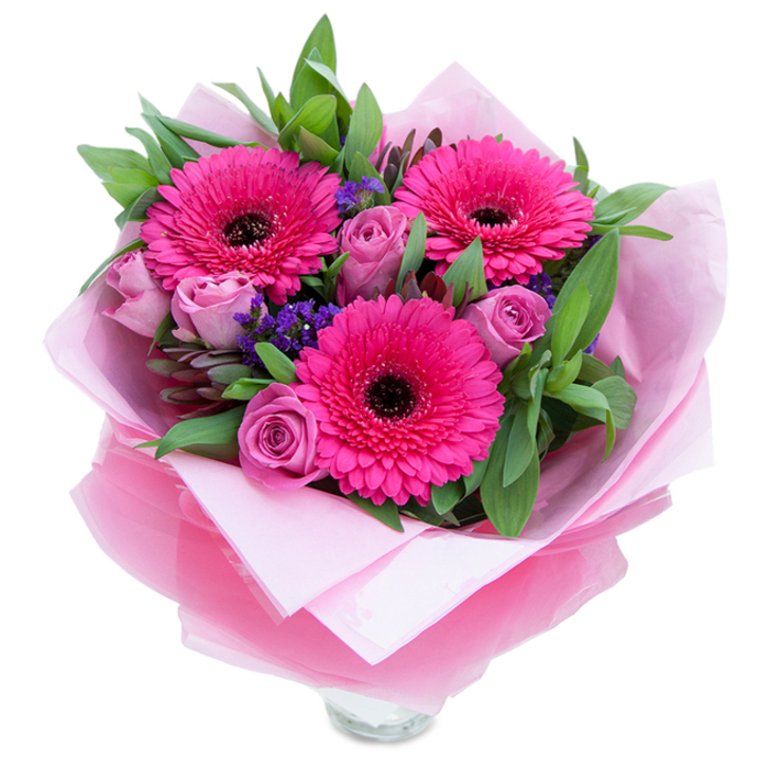 There is a gentle poetry in this arrangement where vivid magenta meets soft lavender. Three fuchsia Gerbera daisies bloom like small suns at the heart of the piece, their concentric petals creating rhythmic movement and a bold visual centrepoint with rich, dark middles. Nestling close, lavender-pink roses unfurl with measured grace-petals layered like the pages of a quiet letter-inviting a closer look and offering a soft, powdery scent. Sprays of purple statice weave through the composition, their tiny flowers bringing an airy, meadow-like texture that contrasts the larger forms. A generous sweep of green foliage - broad salal and aromatic eucalyptus - frames the bouquet, providing both structure and a cool, verdant foil to the warm pinks. The blooms are wrapped in an inner layer of soft matte pale pink tissue and a top layer of translucent fuchsia-pink cellophane that catches the light with a subtle shimmer; the wrapping is folded with intent so the arrangement feels both intimate and refined. The verifiable rim of a clear glass vase sits at the base, a practical cue that this is ready for immediate display. In my mind this bouquet belongs on a mantel in a Farnborough family home after a walk through the town's parks or following the excitement of the airshow - a tactile, fragrant way to celebrate birthdays, anniversaries, or a heartfelt thank-you, crafted with the kind of careful attention you expect from a local florist.