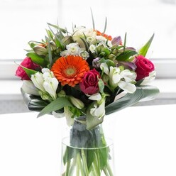 This elegant bouquet is a testament to the balance of style, emotion, and local flavour that defines Flowers Farnborough. Soft, creamy peach and bold orange gerberas form the bouquet's radiant centre, conjuring images of gentle dawns and vibrant sunsets seen from Farnborough's open spaces. Substantial fuchsia roses nestle nearby, their lush, spiralled forms imparting a sense of luxury and timeless affection. Intermingling white alstroemeria and tiny, nuanced filler blooms illuminate the composition, their presence like scattered pearls that reflect the light pouring softly through a sunlit window. Large, glossy green leaves-some broad, others slender-curl around the arrangement, enclosing the mix of hues in a tranquil embrace and lending robust structure. Delicate sprigs of secondary greenery add subtle texture, ensuring every angle of the bouquet appears full and dynamic. Resting in a clear, graceful glass vase, the stems are exposed, providing a connection to the earth and the hands that gathered them. The overall effect is uplifting and expressive, a fitting centrepiece for community gatherings in Farnborough or cherished family celebrations. Each element is handled with care and artistry, shining with freshness and designed to evoke hope and happiness.